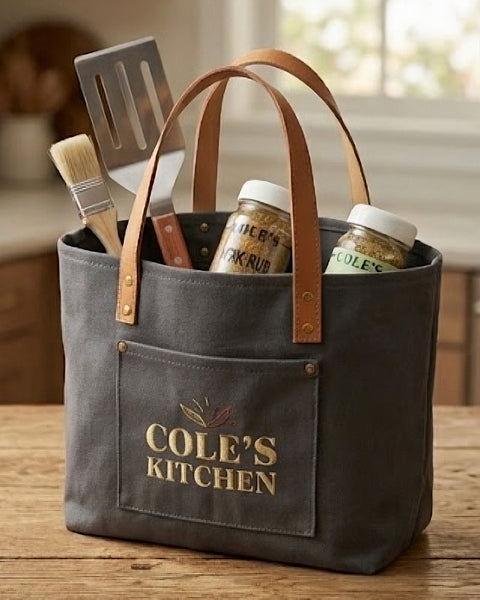 A durable dark gray canvas kitchen tote bag with tan leather handles sits on a wooden table, holding kitchen tools and botanical small-batch spice jars. The front pocket is embroidered by Northern Loom with 'COLE'S KITCHEN' branded in high-density gold lettering.