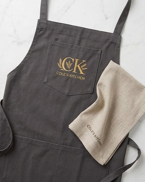 A Northern Loom dark charcoal gray kitchen apron with high-density gold embroidered 'CK Cole's Kitchen' logo on the chest pocket, styled flat-lay with a beige kitchen towel on a white marble background.