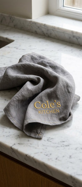 A Northern Loom gray linen kitchen towel with custom brand logo embroidered text reading 'Cole's Kitchen' resting on a white marble countertop near a window.