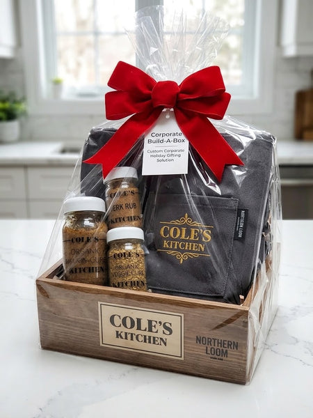 A custom gift box set from Cole's Kitchen and Northern Loom, wrapped in clear cellophane with a large red bow. The set includes a wooden box labeled 'COLE'S KITCHEN', a black apron with embroidered gold lettering by Northern Loom, organic jerk marinade and two artisanal spice blends.