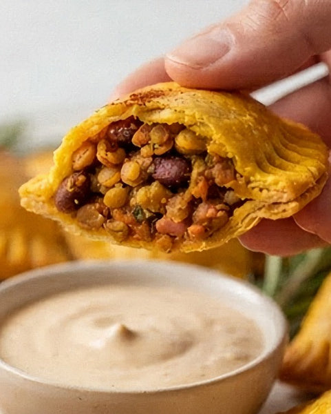 A hand holds a flaky golden yellow baked Jamaican patty stuffed with a colourful lentil and vegetable mixture, with a small bowl of creamy beige dipping sauce in the foreground.