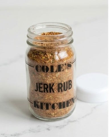 A clear glass mason jar filled with a coarse brown with green-accents seasoning rub, labeled 'COLE'S JERK RUB KITCHEN' in black text, with a white lid resting beside it on a light marble surface.