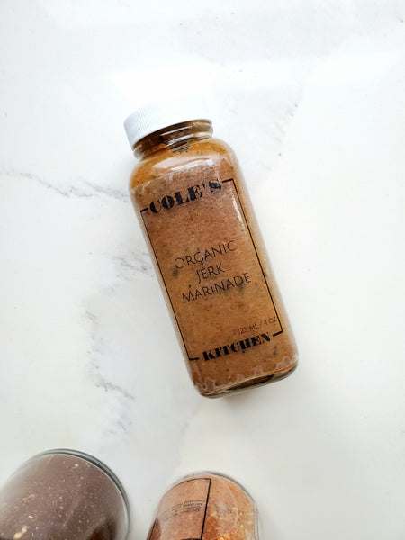A tall clear glass bottle of Cole's Kitchen Organic Jerk Marinade showing the rich brown texture of the spices, placed on a white marble surface.
