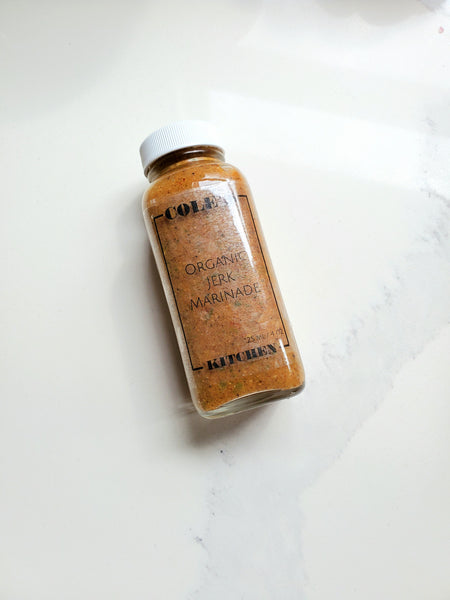 A small square glass jar of Cole's Kitchen Organic Jerk Marinade with a white shaker lid, showing the coarse spice blend inside, on a clean white surface.