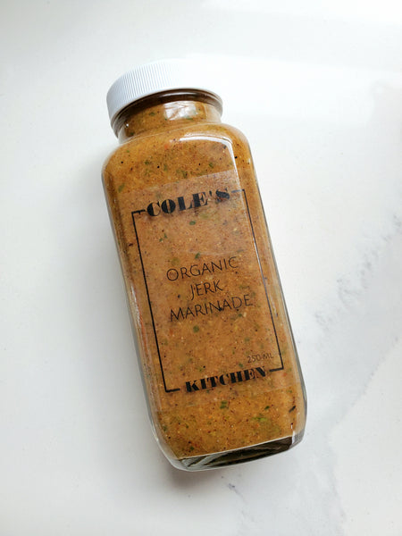 A glass jar of Cole's Kitchen Organic Jerk Marinade with a white lid, showcasing the textured green and brown herb-and-spice blend on a marble background.