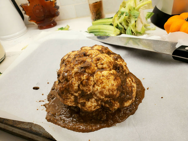 A whole roasted cauliflower head smothered in rich, dark Cole's Kitchen Organic Jerk Marinade on parchment paper, with fresh kitchen ingredients in the background.