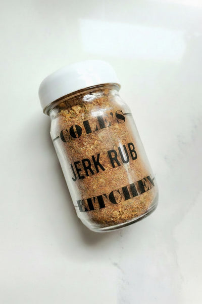 A small square glass jar of Cole's Kitchen Organic Jerk Spice Rub with a white lid, angled to show the coarse blend of Caribbean spices on a white background