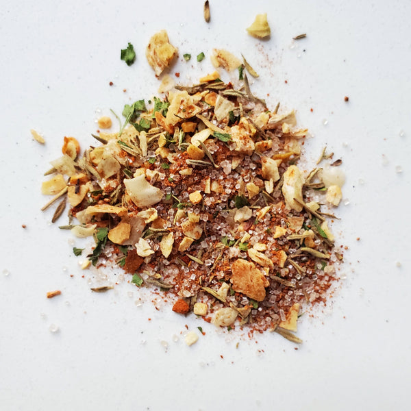 A close-up overhead view of Cole's Kitchen Organic Jerk Spice Rub scattered on a white surface, showing the coarse texture of thyme, chili peppers, and aromatic Caribbean spices.