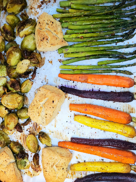 A sheet pan of roasted vegetables including carrots, asparagus, and brussels sprouts seasoned with Cole's Kitchen Veggie Cure Spice Blend, served alongside roasted chicken or tofu.