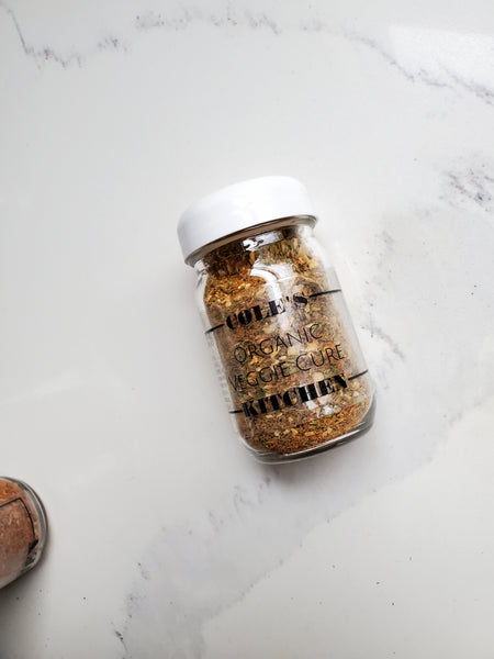 A 4 oz glass jar of Cole's Kitchen Organic Veggie Cure Spice Blend with a white lid, positioned on a white marble surface to show the colorful herb and spice layers.