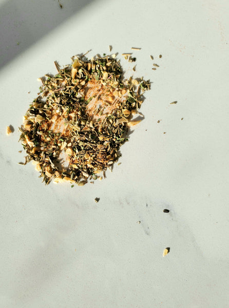 A close-up overhead view of the Veggie Cure Spice Blend scattered on a white surface, showing the mix of dried green herbs like thyme and oregano with ground spices.